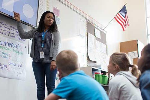 person pointing a screen displaying shapes while children sitting look on